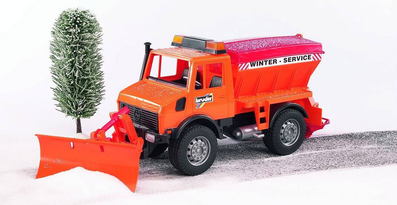 Ein rotes Spielzeug-Schneepflugfahrzeug mit der Aufschrift 'Winter-Service' fährt auf einer schneebedeckten Straße neben einem Baum.