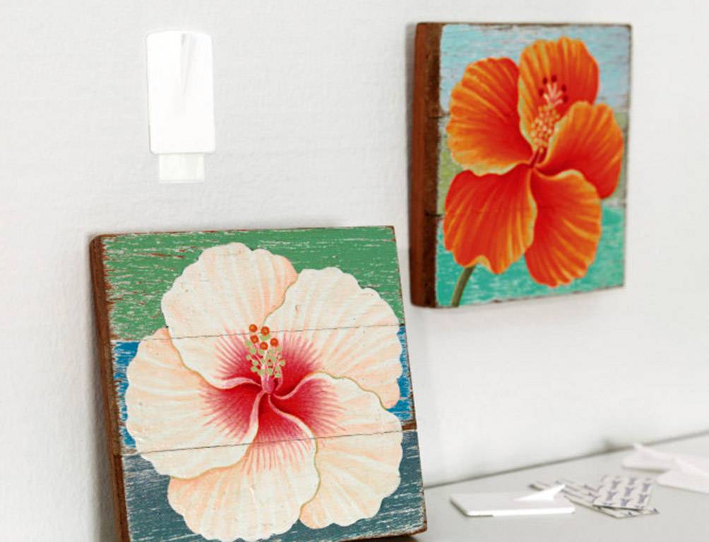 Zwei Holzbilder von Hibiskusblüten in kräftigen Farben hängen an einer hellen Wand. Links oben ist ein kleiner, weißer Lufterfrischer.