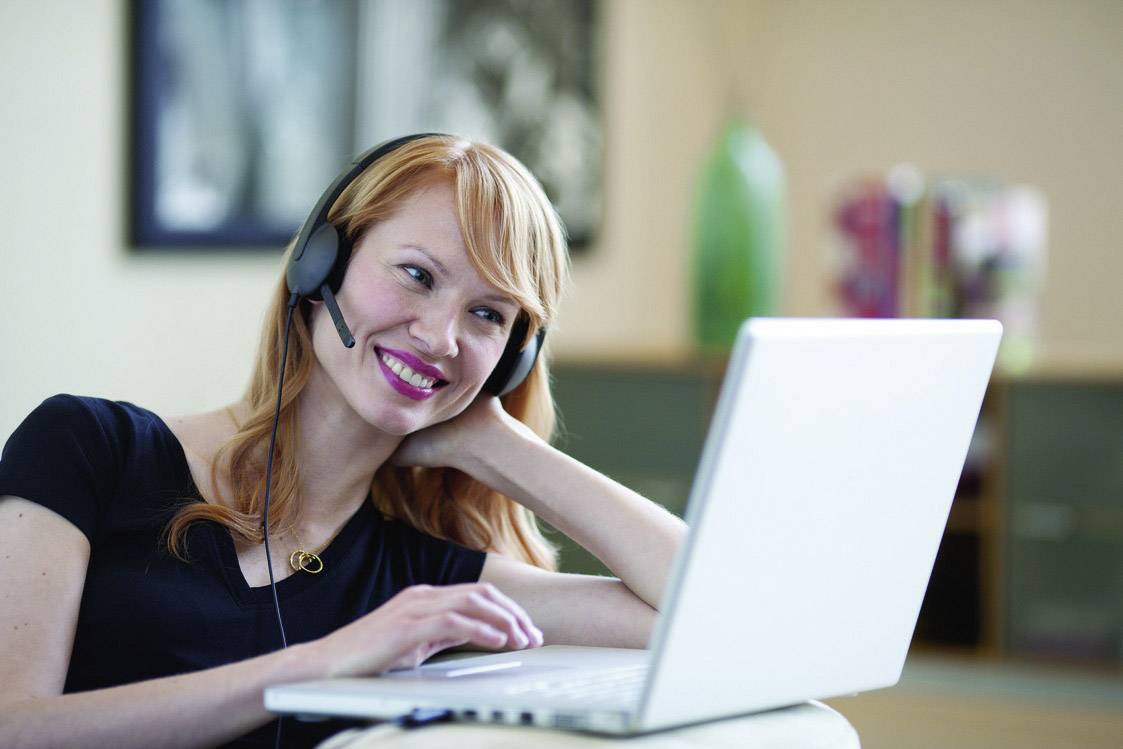 Eine lächelnde Person mit Headset sitzt vor einem Laptop und scheint an einem virtuellen Meeting teilzunehmen.
