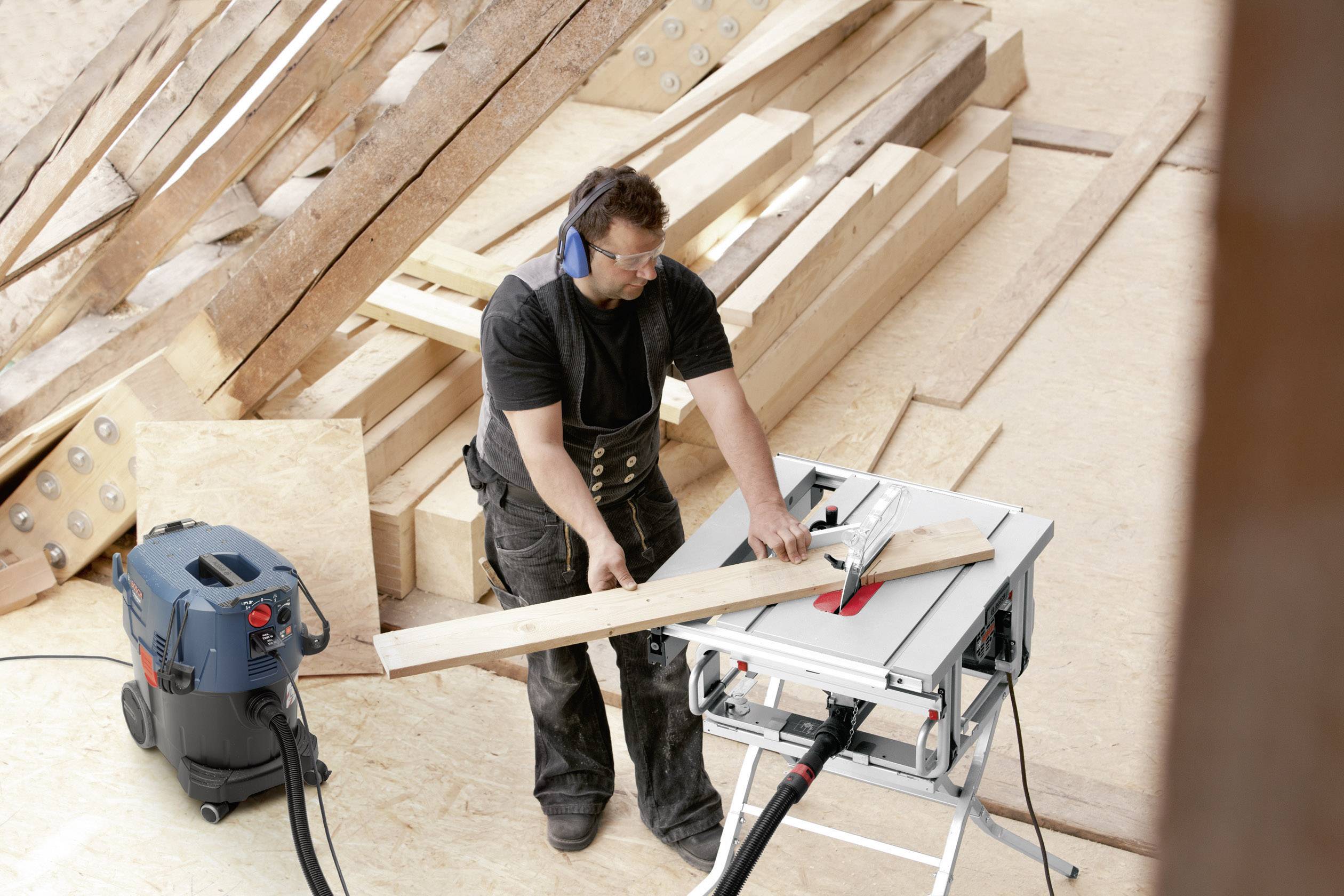 Ein Mann sägt mit Gehörschutz und Schutzbrille ein Holzbrett auf einer Baustelle. Er verwendet eine Tischkreissäge, um präzise Schnitte auszuführen.