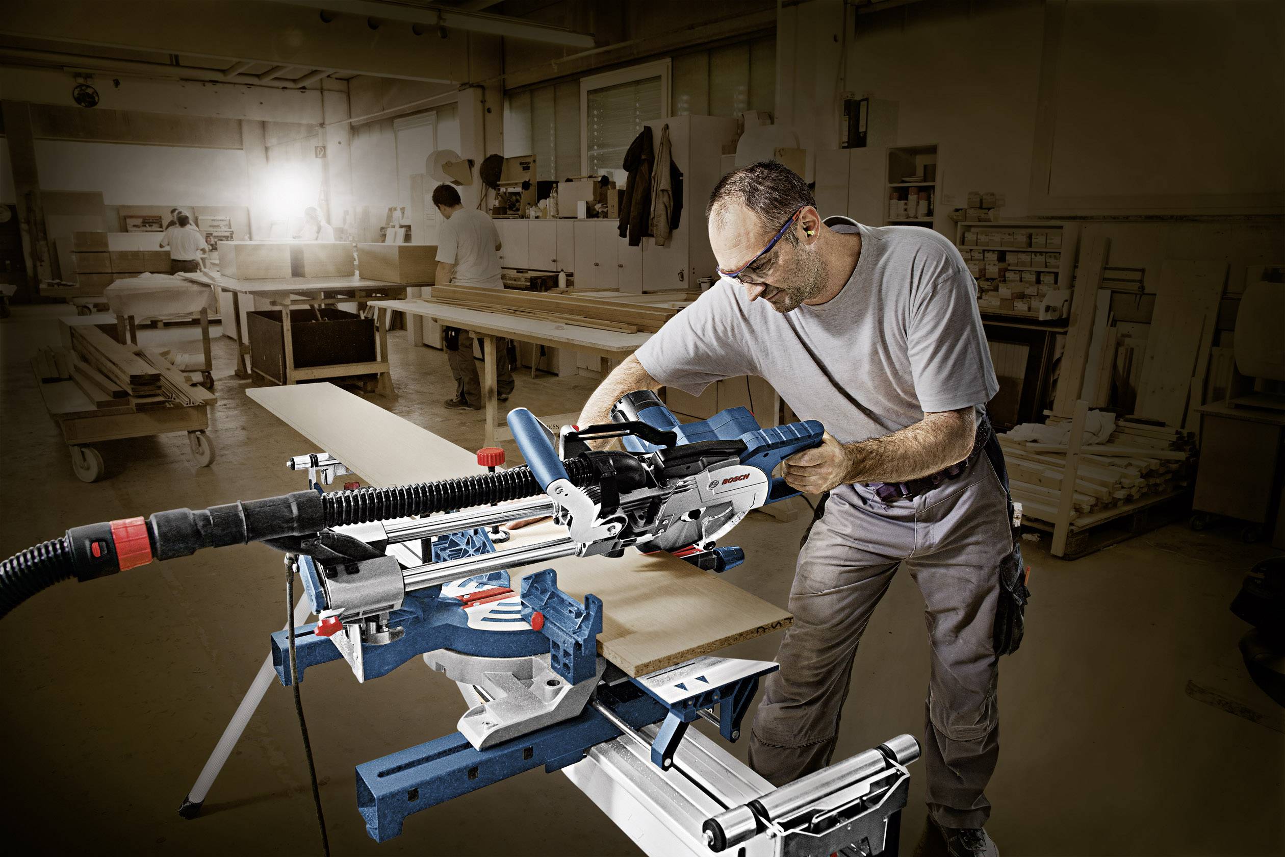 Ein Handwerker in einer Werkstatt bedient eine Kreissäge. Er trägt Schutzbrillen und konzentriert sich auf die Arbeit an einem Holzstück. Im Hintergrund sind Werkbänke und Werkzeuge sichtbar.