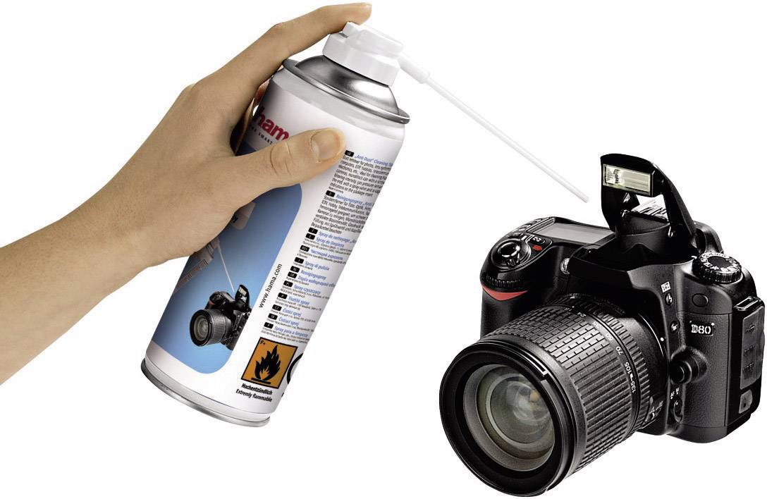 Eine Hand hält eine Druckluftdose nahe einer Kamera, um Staub oder Schmutz von deren Sensor oder Gehäuse zu entfernen.