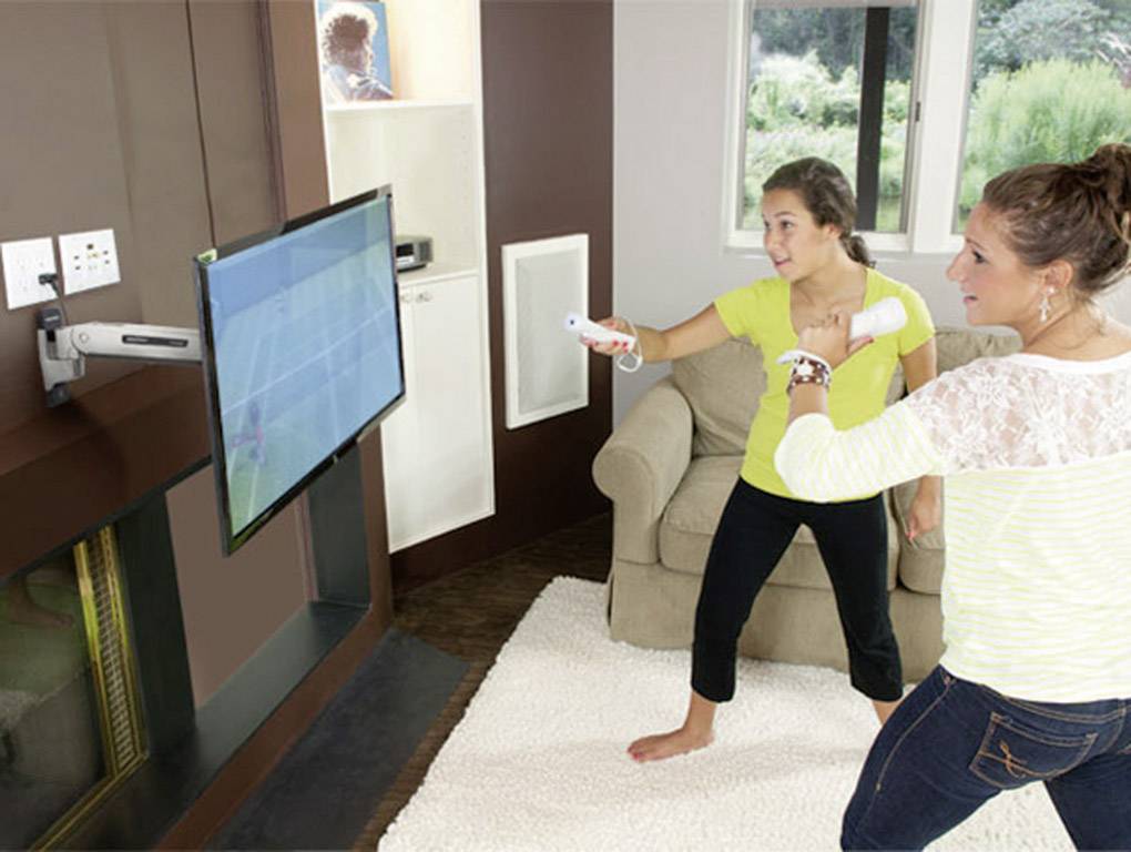 Zwei Personen spielen ein Tennisspiel auf einer Spielkonsole in einem Wohnzimmer, konzentriert und in Bewegung vor dem Bildschirm.