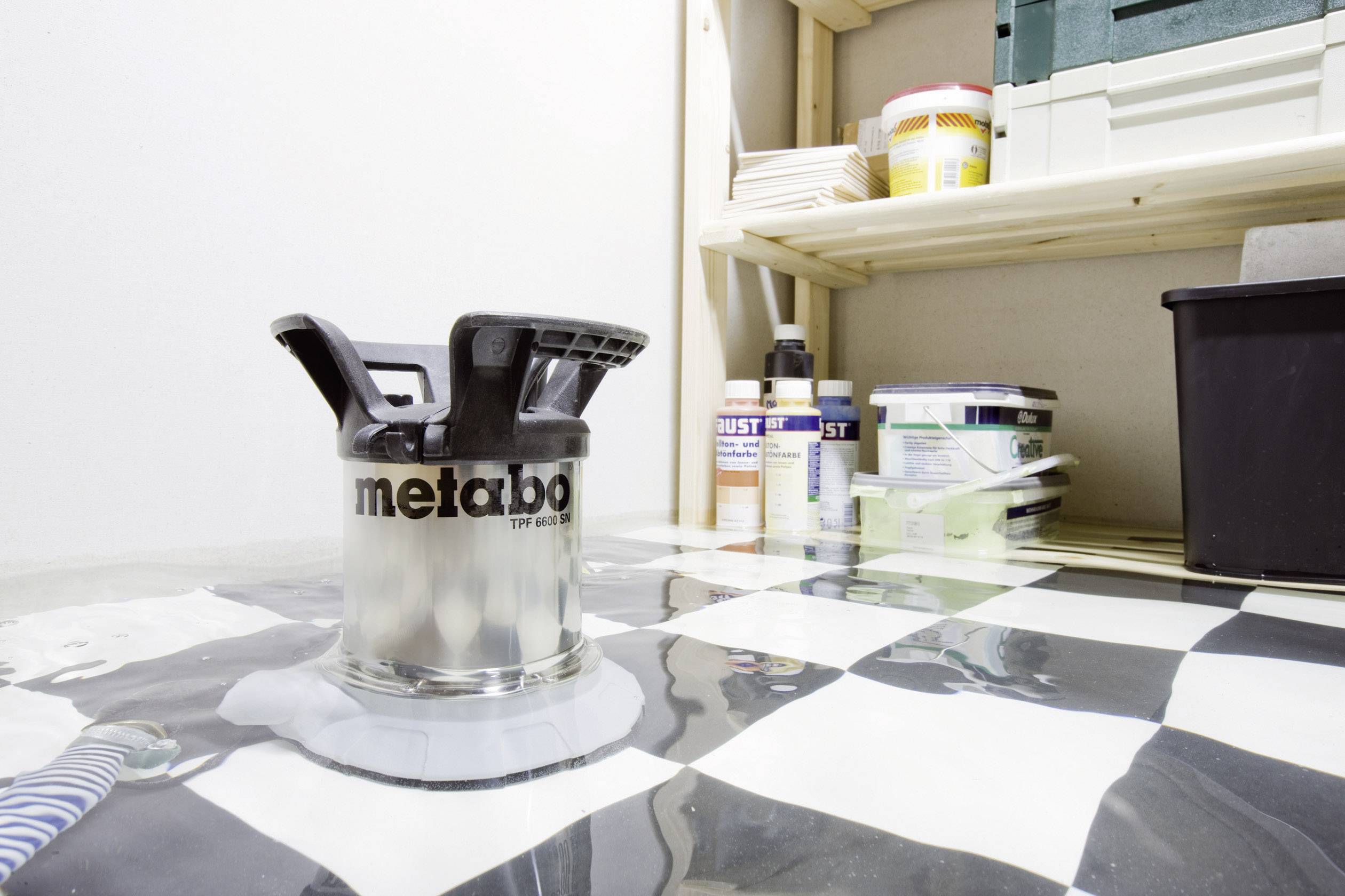 Ein Wasserabsauger wird auf einem gefluteten Schachbrettboden in einem Keller genutzt, um Wasser abzupumpen. Im Hintergrund sind Regale mit Werkzeugen.