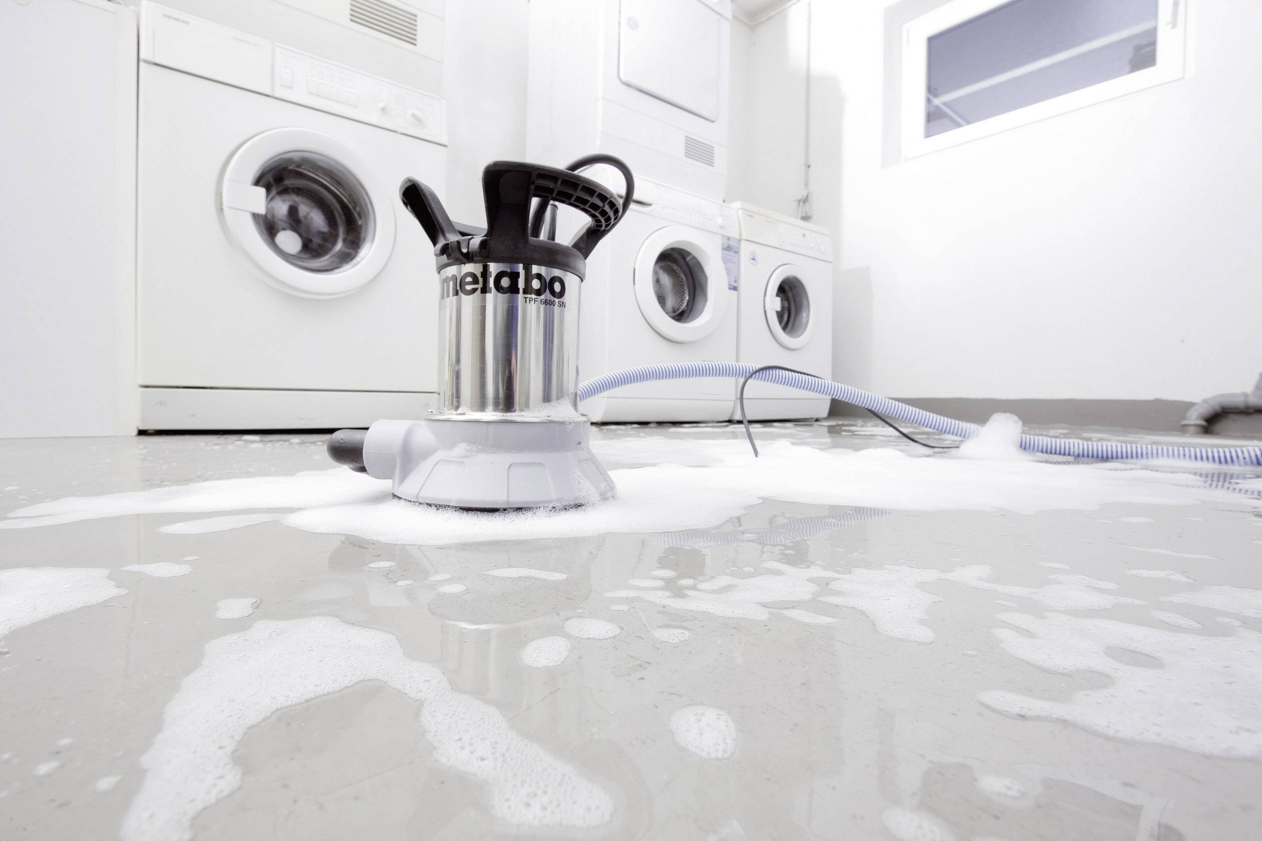 Eine Waschmaschine in einem weißen Raum hat übergelaufen; Wasser und Schaum bedecken den Boden, eine Pumpe steht bereit zum Abpumpen.