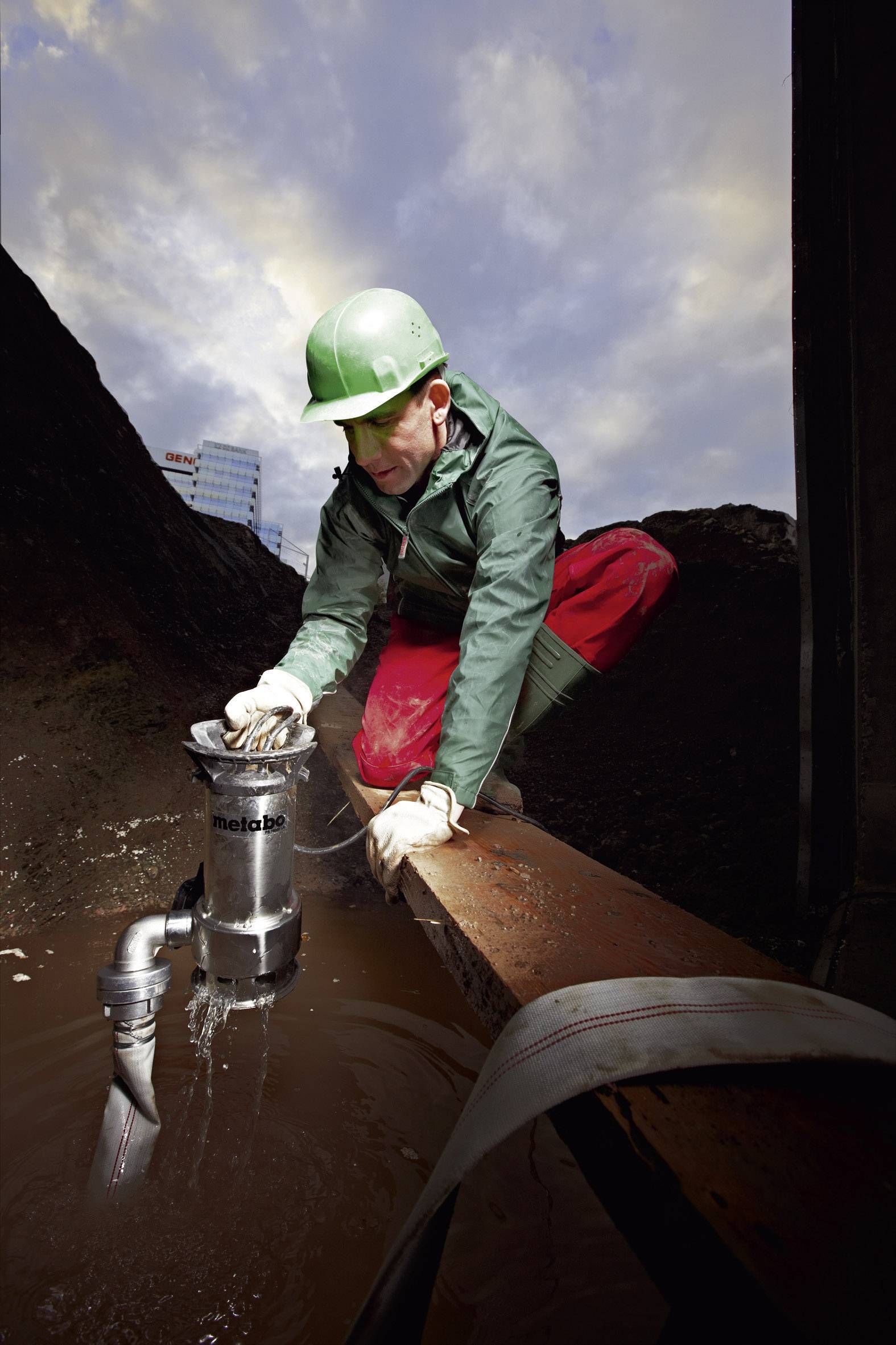Ein Bauarbeiter mit grünem Helm installiert eine Pumpe in einem schlammigen Bereich auf einer Baustelle bei bewölktem Himmel.