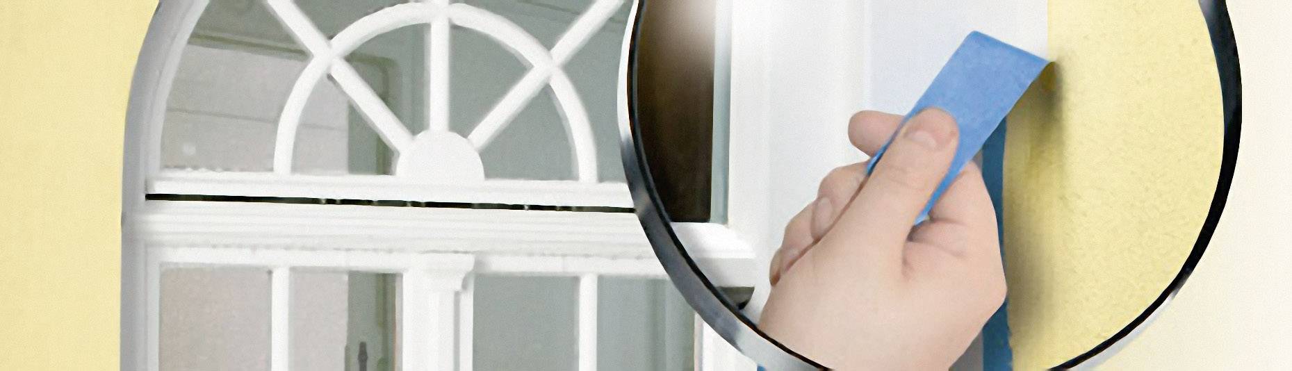 Eine Hand klebt blaues Malerband um das Fenster einer gelben Wand ab, um es vor Farbspritzern zu schützen.