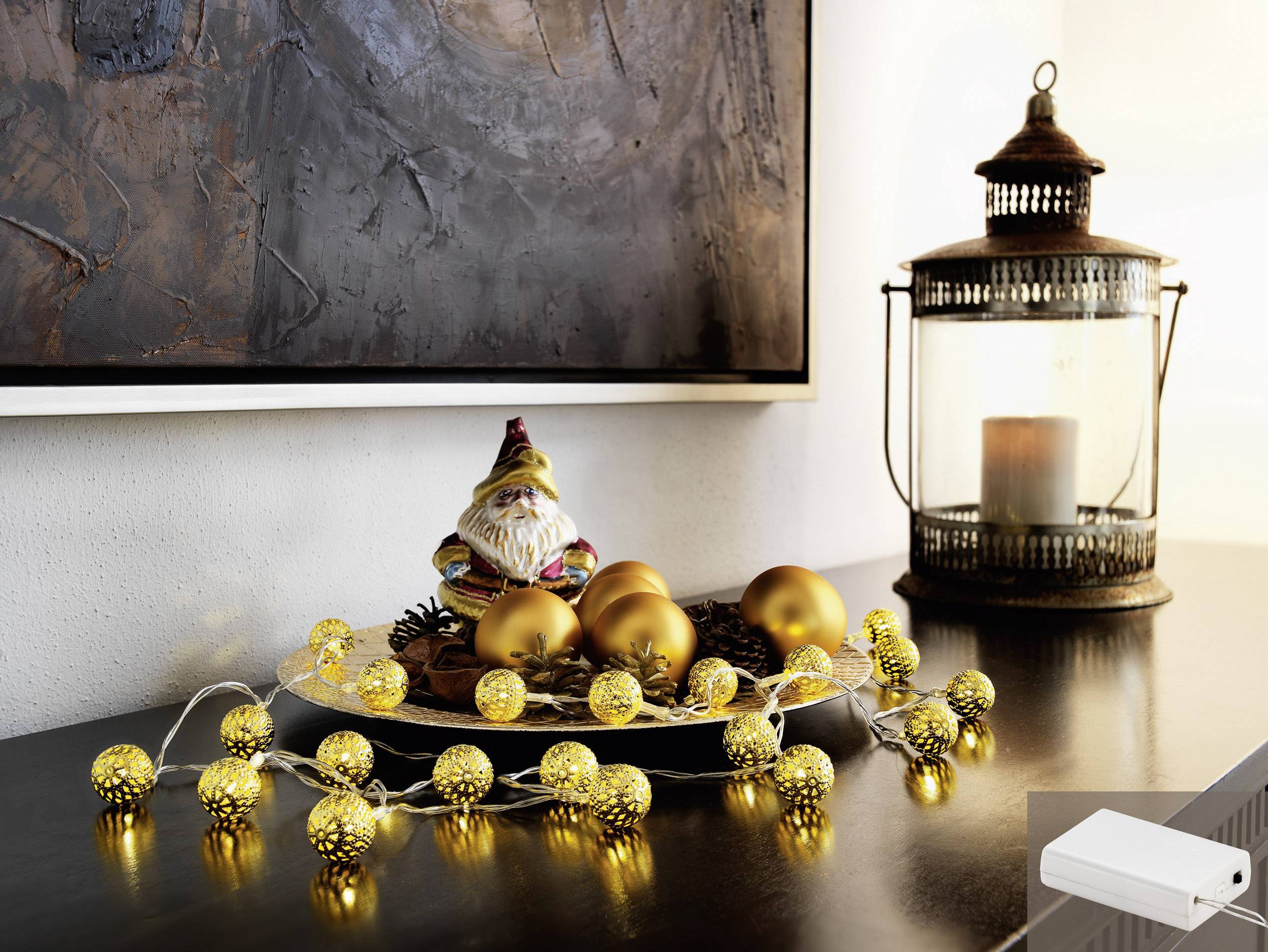 Ein dekorativer Tisch mit goldenen Weihnachtskugeln und Lichterkette, daneben eine Laterne und eine kleine Weihnachtsfigur.