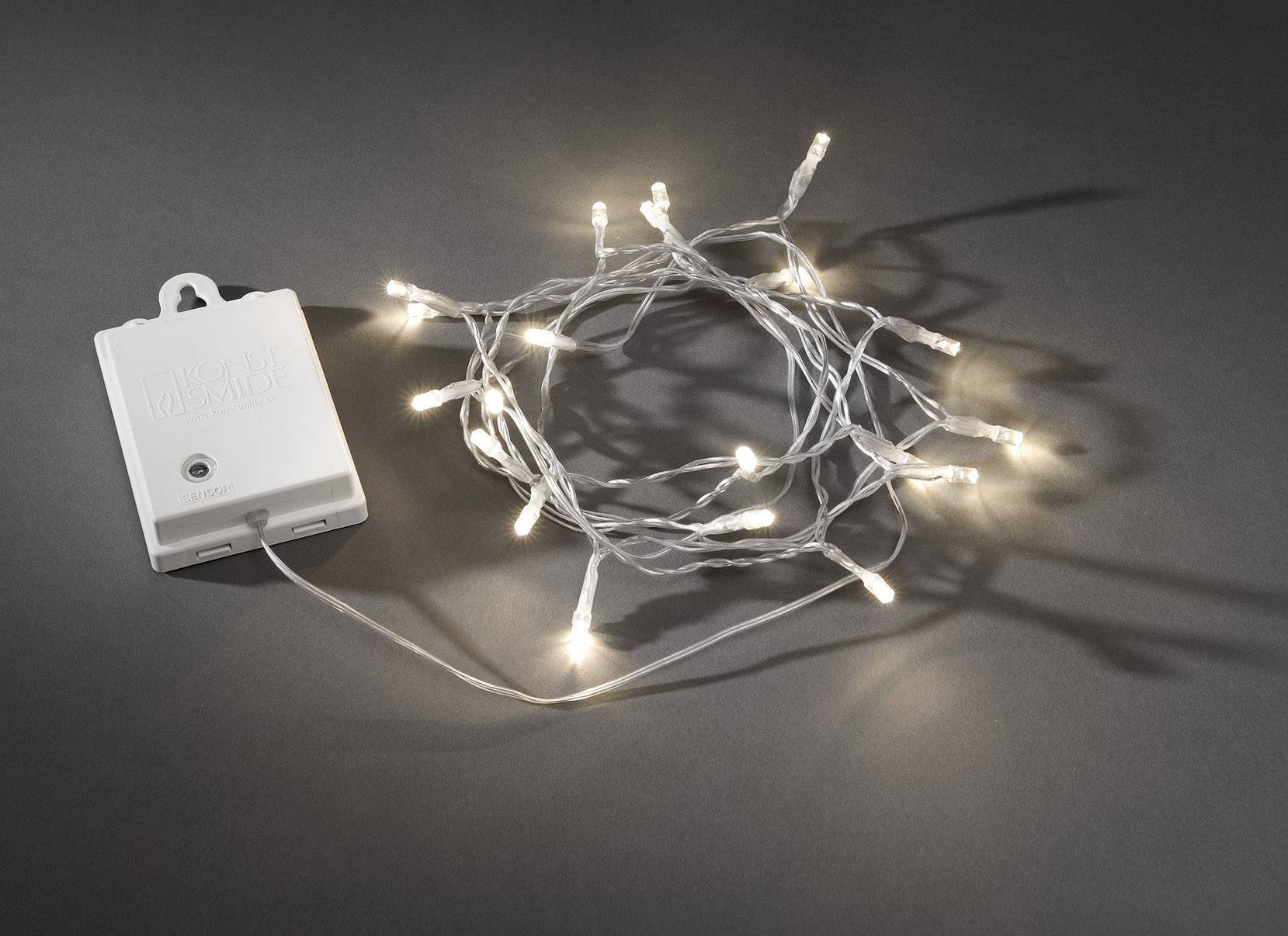 Eine leuchtende Lichterkette mit weißem Kabel ist neben einer weißen, batteriebetriebenen Steuerbox auf einem dunklen Hintergrund abgebildet.