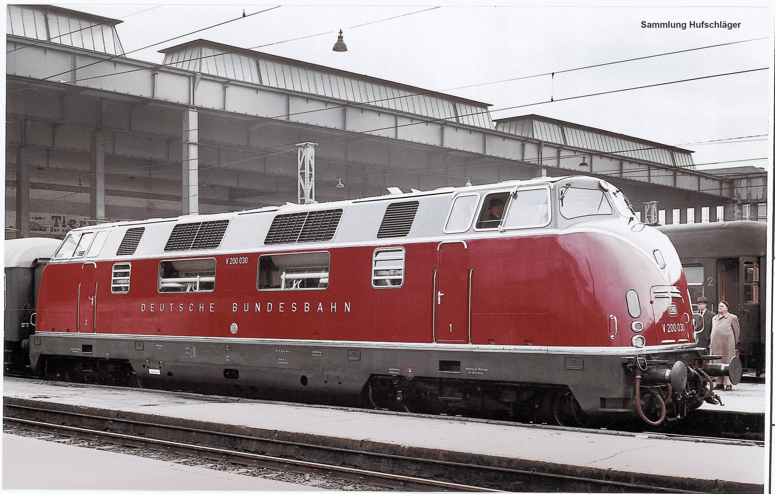 Ein roter Diesellokomotiven-Zug mit der Aufschrift 'Deutsche Bundesbahn' steht auf einem Bahnhofsgleis. Gebäude und Personen im Hintergrund.