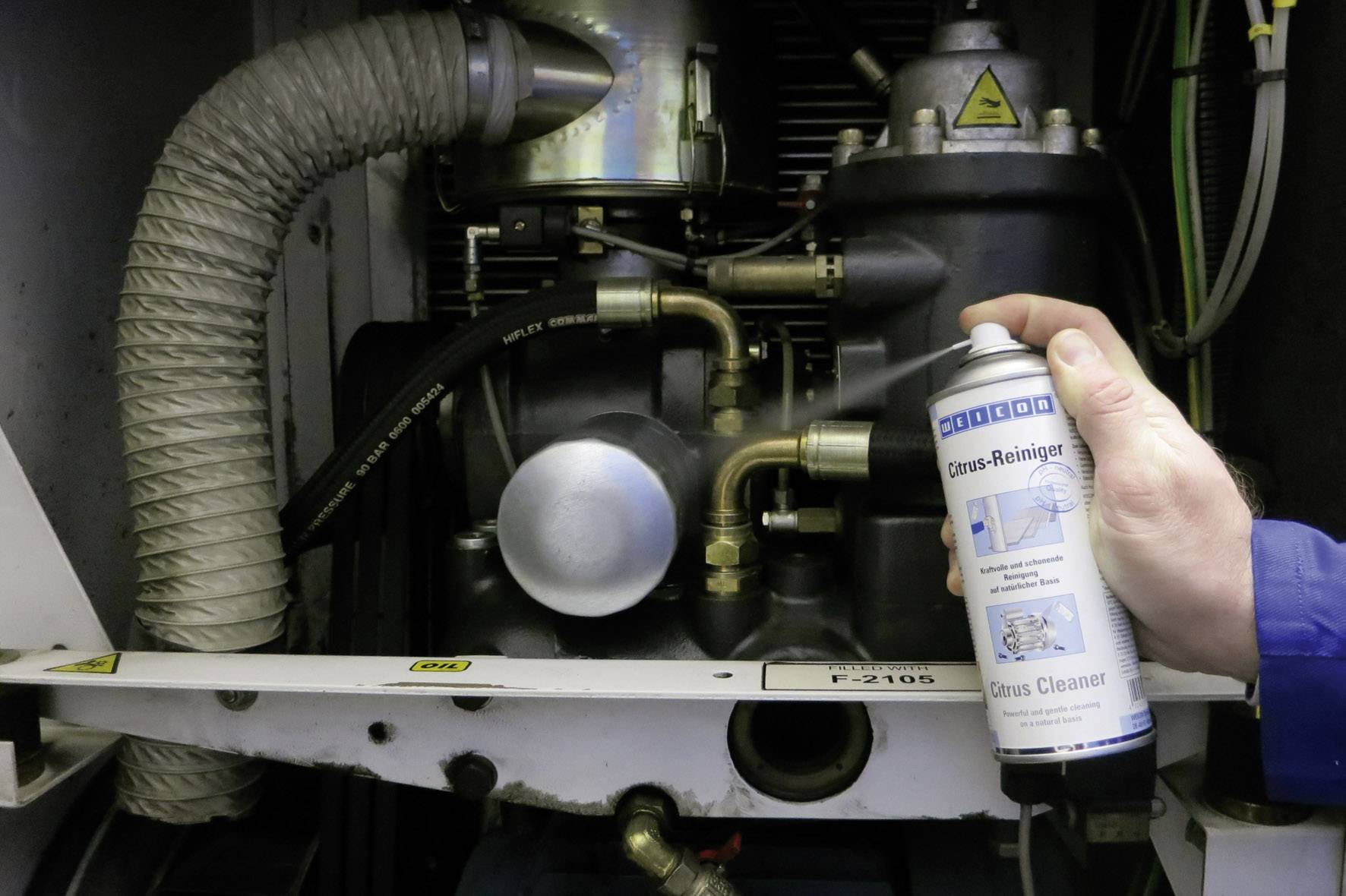 Eine Person hält eine Dose Gasreiniger vor einer Maschine mit Schläuchen und Rohren. Die Szene zeigt den Reinigungsprozess.
