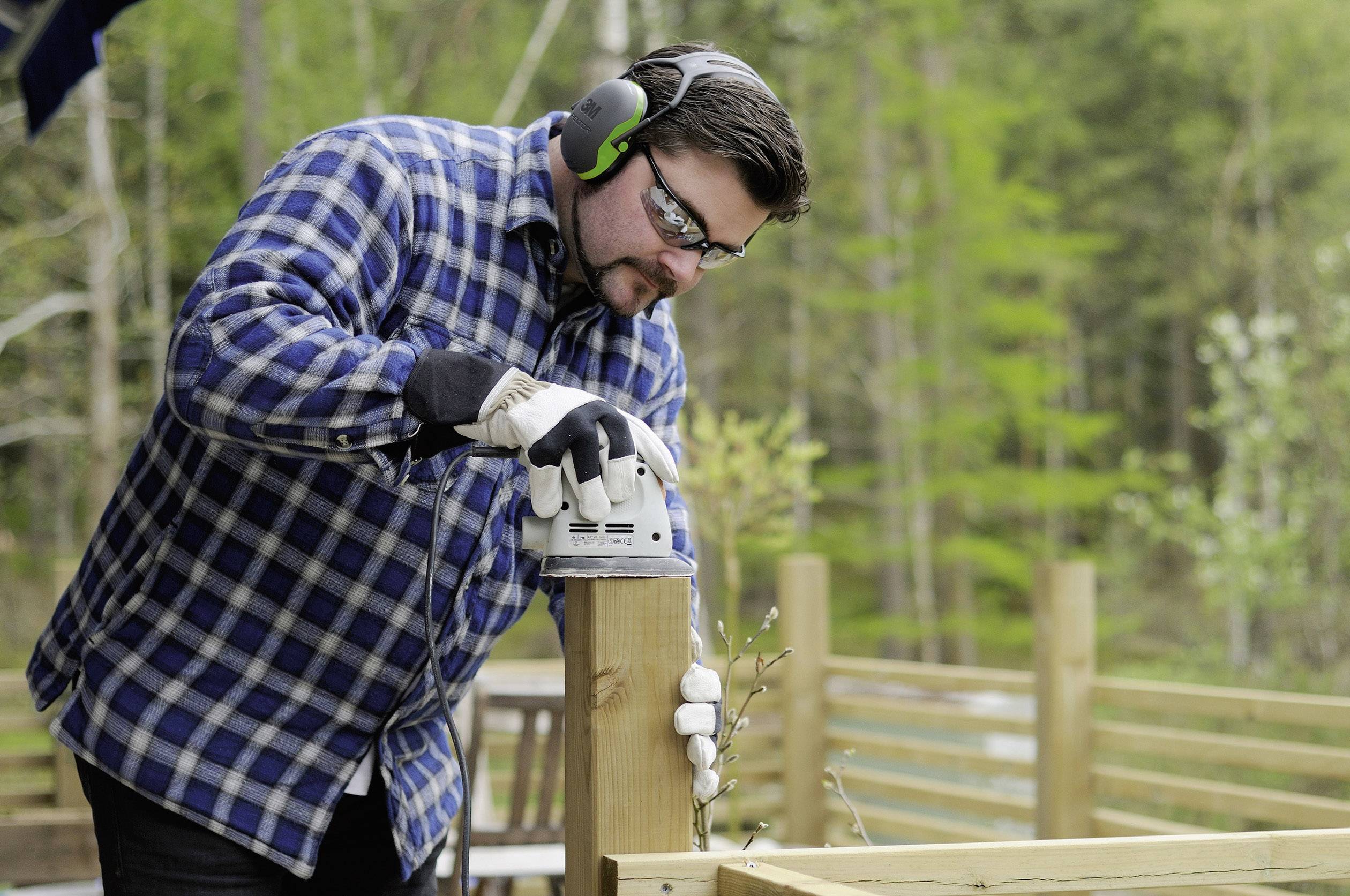 Ein Mann arbeitet mit einer elektrischen Schleifmaschine auf einer Holzterrasse, trägt Schutzbrille und Gehörschutz, umgeben von Wald.