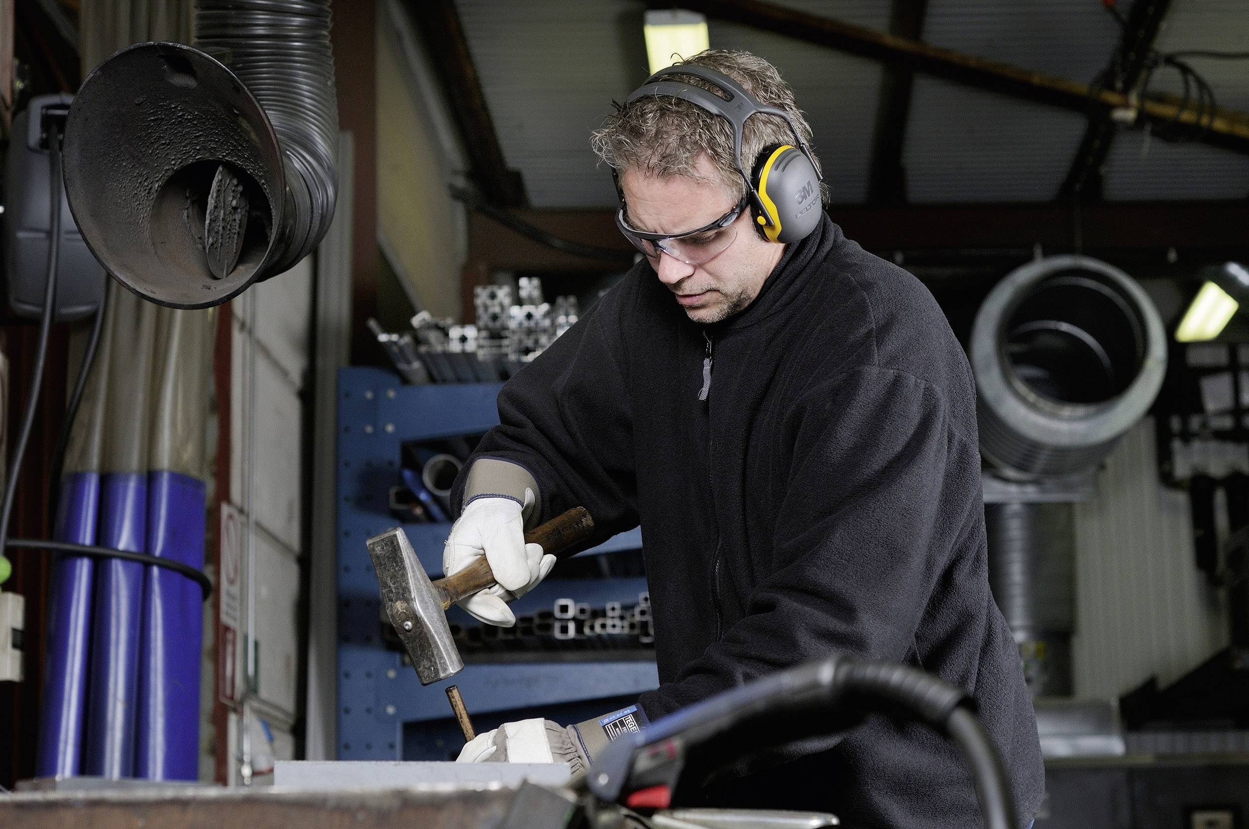 Ein Mann in Schutzkleidung und Gehörschutz arbeitet in einer Werkstatt mit einem Hammer an einem Metallstück. Im Hintergrund sind Regale mit Material zu sehen.