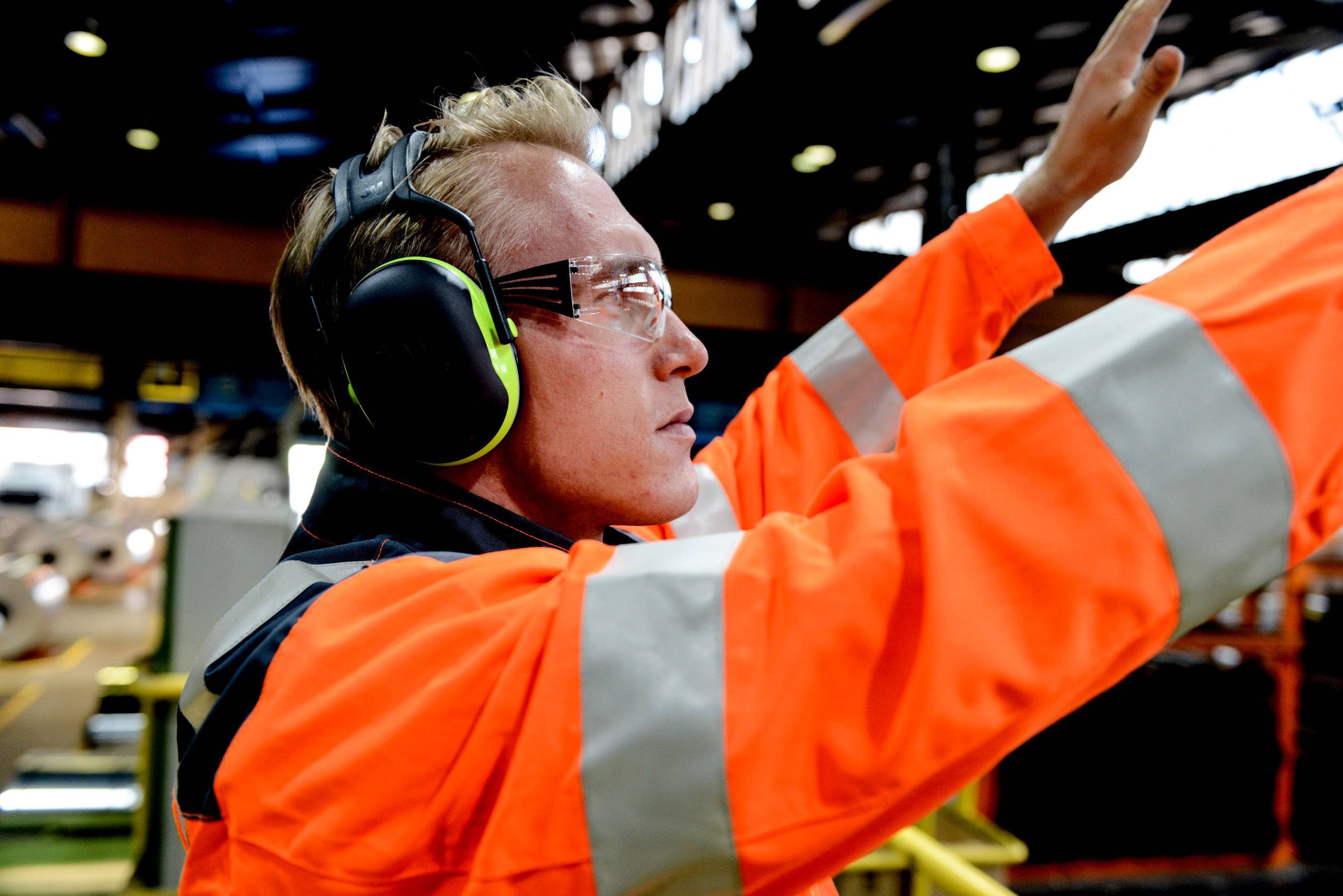 Ein Arbeiter in orangener Schutzkleidung und Gehörschutz hebt einen Arm, möglicherweise um ein Signal in einer Fabrikumgebung zu geben.