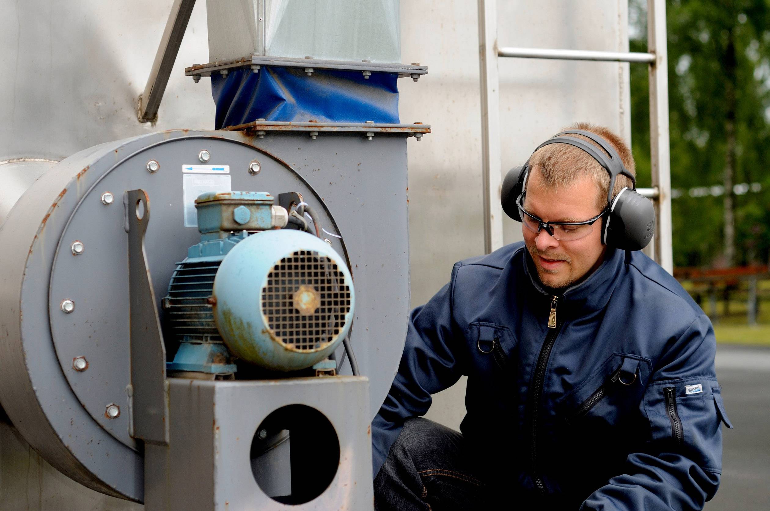 Ein Mann trägt Ohrenschützer und Schutzbrille, während er eine große Maschine im Freien inspiziert.