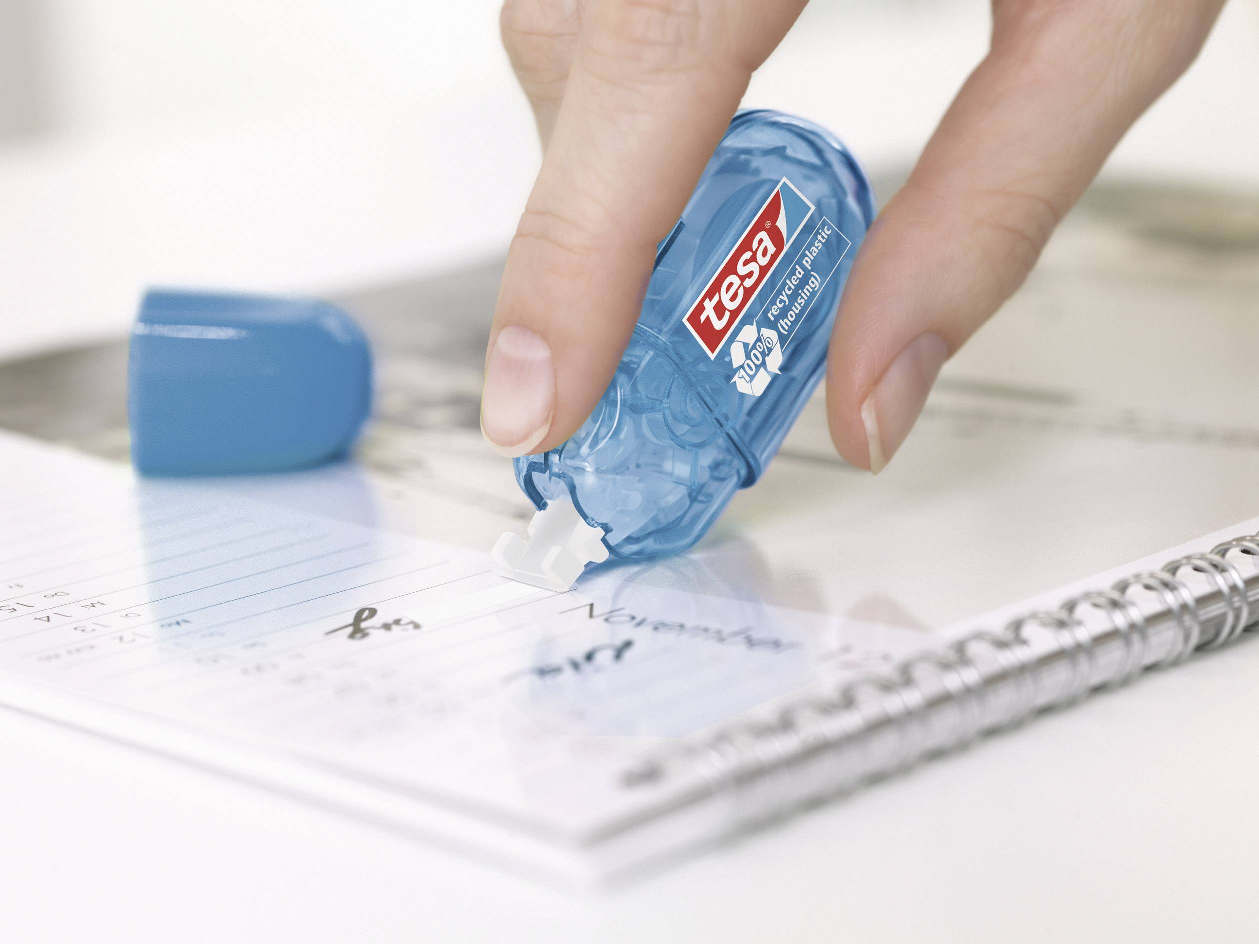 Eine Hand verwendet einen blauen Korrekturroller, um Fehler auf einem Notizbuch zu korrigieren. Der Roller trägt die Marke 'tesa'.