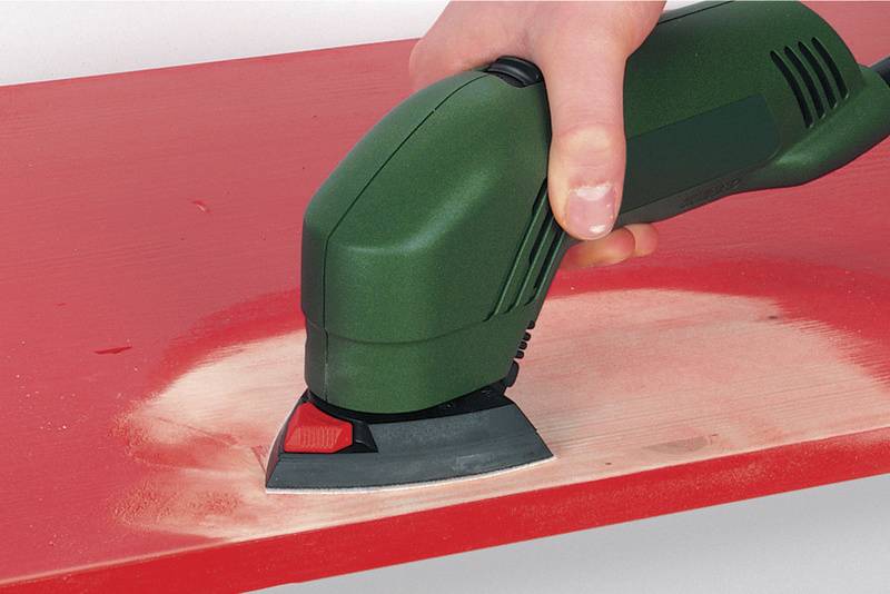 Eine grüne handgehaltene Schleifmaschine schleift eine rote Holzoberfläche ab. Staub sammelt sich während des Schleifens.