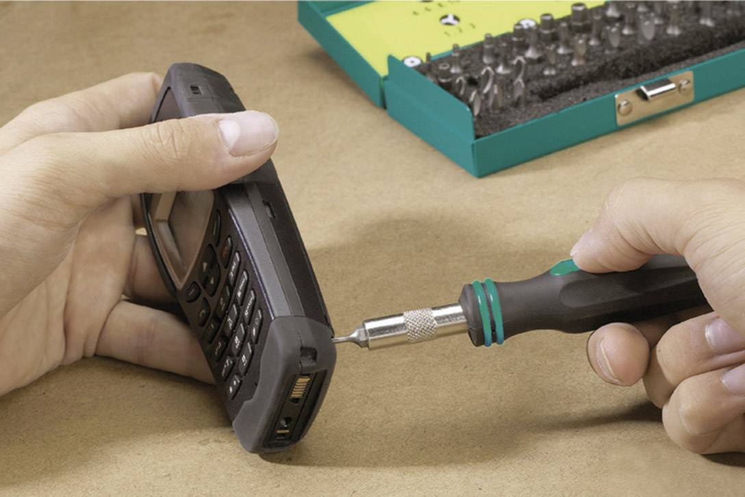 Eine Person öffnet ein Mobiltelefon mit einem Schraubendreher. Im Hintergrund ist ein grüner Werkzeugkasten sichtbar.