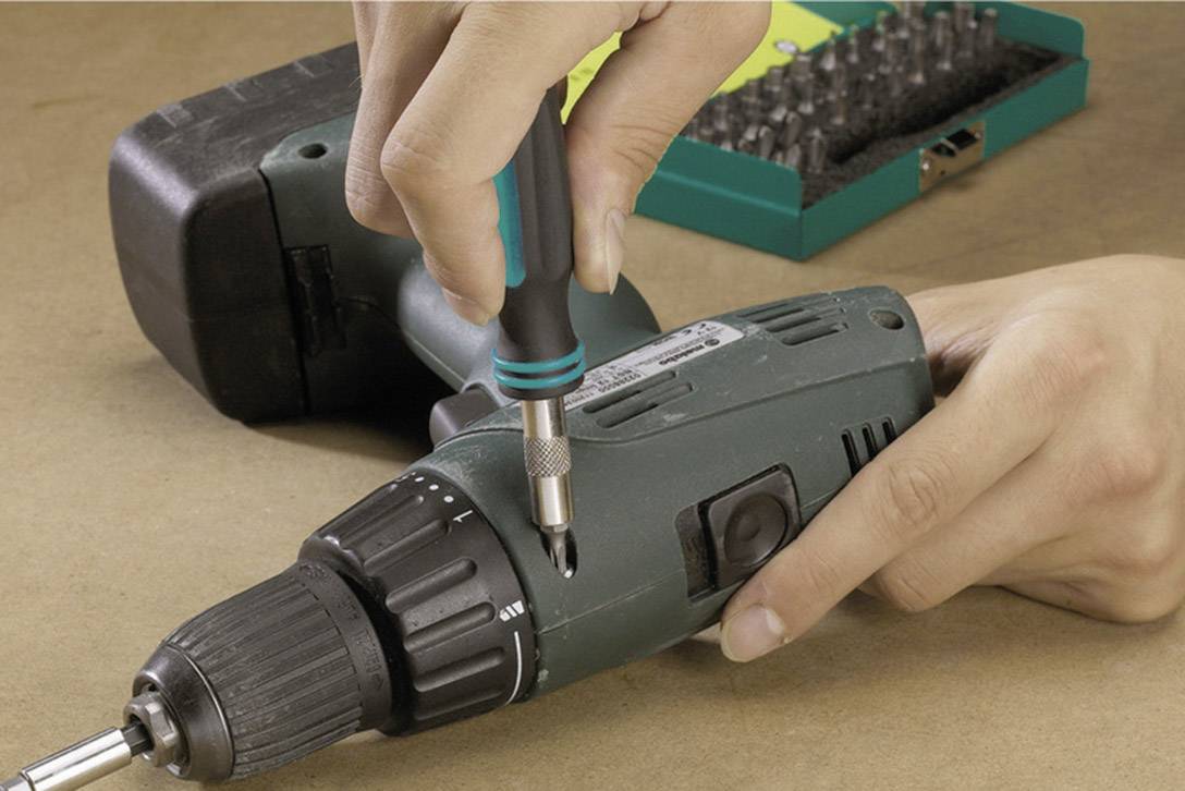 Person hält einen Schraubendreher und schraubt an einem Akkuschrauber. Im Hintergrund ist ein Bit-Set zu sehen.