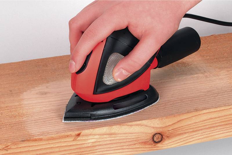 Eine Hand hält einen roten Deltaschleifer, der eine Holzoberfläche schleift. Das Holz zeigt bereits bearbeitete und unbearbeitete Bereiche.