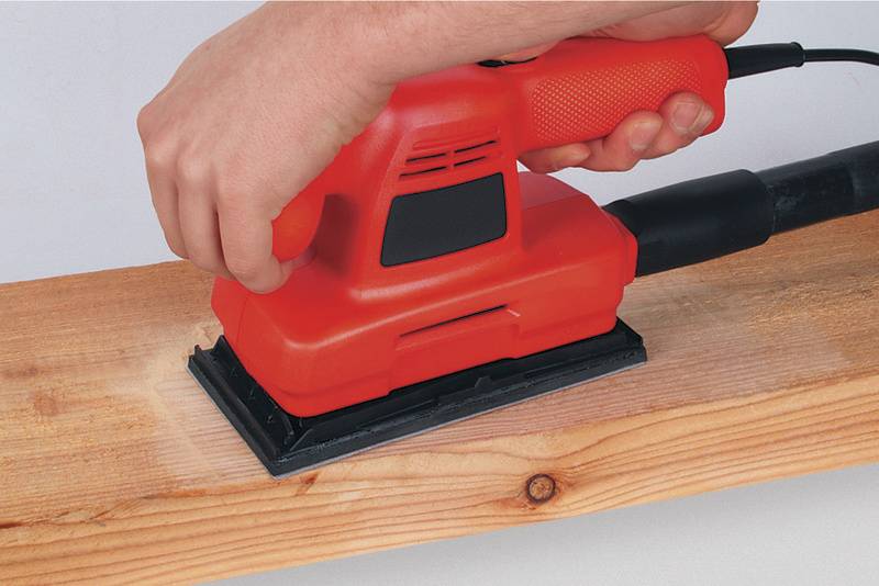 Eine Hand hält einen roten Schwingschleifer, der über ein Holzbrett geführt wird, um dessen Oberfläche zu glätten.