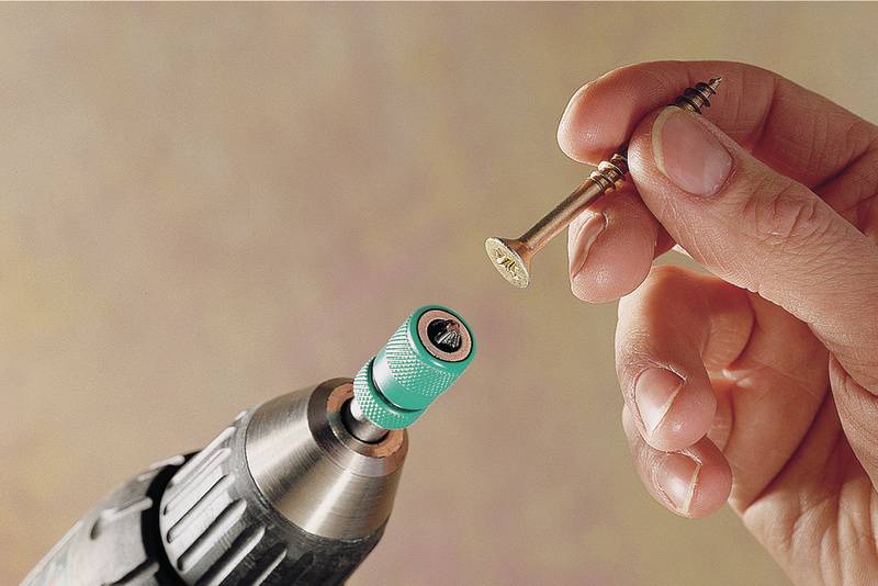 Eine Hand hält eine Schraube vor einem Akkuschrauber mit grünem Bit. Vorbereitung zum Einschrauben in eine Oberfläche.