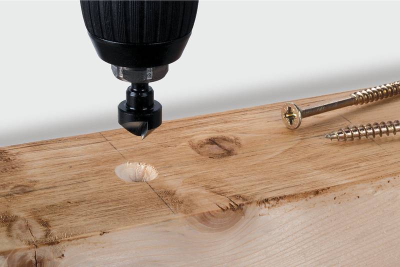 Ein Holzbrett mit einem Bohrloch wird von einem Bohrer bearbeitet. Zwei Schrauben liegen daneben.