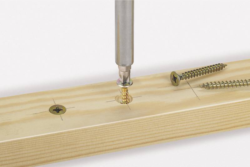 Ein Schraubendreher zieht eine goldene Schraube in ein Holzbrett ein. Zwei weitere Schrauben sind auf dem Brett sichtbar.