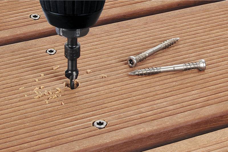 Nahaufnahme eines Bohrers, der eine Schraube in eine Holzplatte dreht. Daneben liegen zwei weitere Schrauben auf der Holzoberfläche.