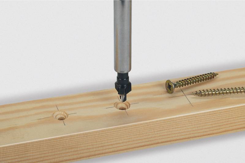 Ein Bohrer senkt Löcher in ein Holzbrett, daneben liegen zwei Holzschrauben.