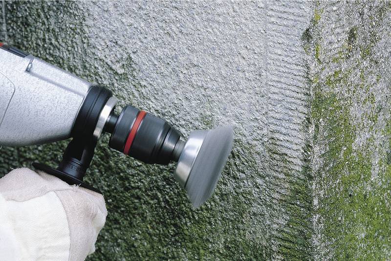 Eine Person bearbeitet mit einem Winkelschleifer eine betonierte Wand, die eine grünliche Patina aufweist.