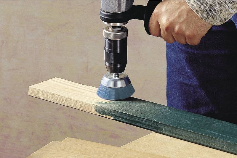 Eine Hand hält eine Bohrmaschine mit einem Drahtbürstenaufsatz, um grüne Farbe von einem Holzbrett abzuschleifen.