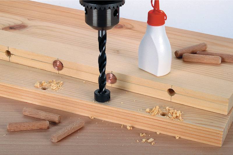 Ein Forstnerbohrer bohrt ein Loch in zwei übereinanderliegende Holzbretter. Umgeben von Dübeln, Holzspänen und einer Leimflasche.