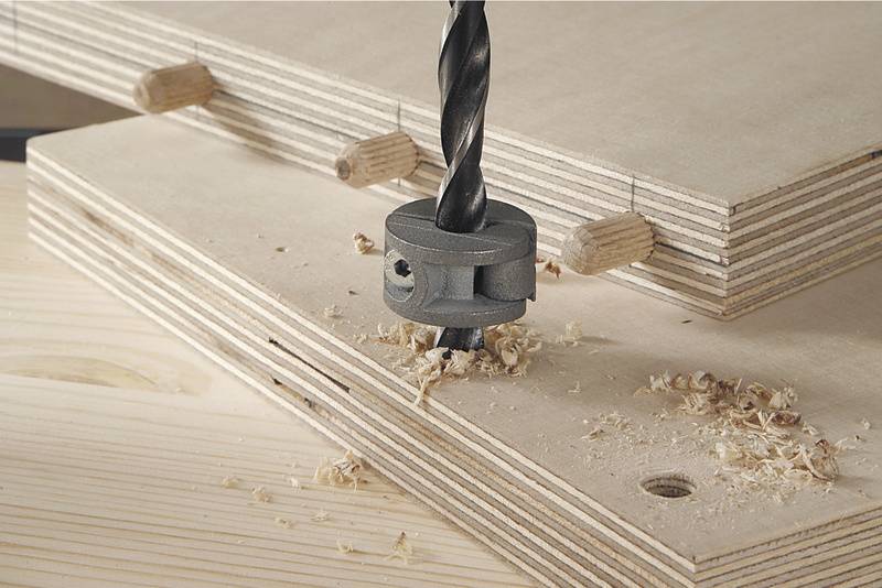 Ein Bohrer mit einem Bohranschlag bohrt ein Loch in zwei übereinanderliegende Holzplatten. Dübel sind zur Verbindung eingesetzt.