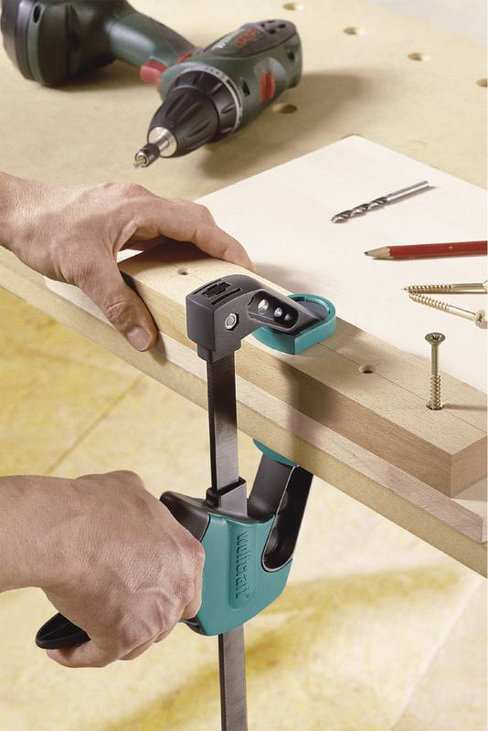 Eine Person befestigt ein Holzbrett mit einer Zwinge an einem Tisch. Auf dem Tisch liegt eine Bohrmaschine, Bleistift und Schrauben.