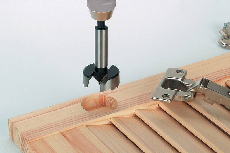 Ein Bohrer erstellt ein Loch in einem Holzbrett, neben einer Scharnierbefestigung.