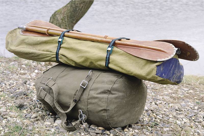 Ein olivgrüner Seesack liegt auf steinigem Boden am Flussufer. Obenauf sind ein gepacktes Zelt und zwei Holzpaddel befestigt.