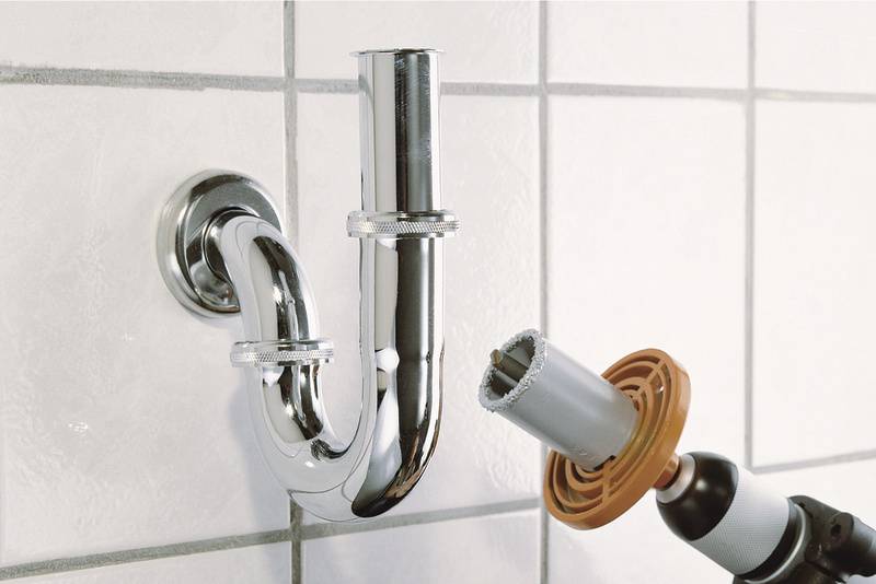 Ein verchromtes Abflussrohr in S-Form wird an einer gefliesten Wand installiert. Eine Bohrmaschine mit einer Lochsäge ist in der Nähe.