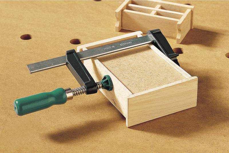 Ein Holzkasten wird mit einer Schraubzwinge auf einem Arbeitstisch fixiert. Im Hintergrund ist eine kleine Holzbox zu sehen.