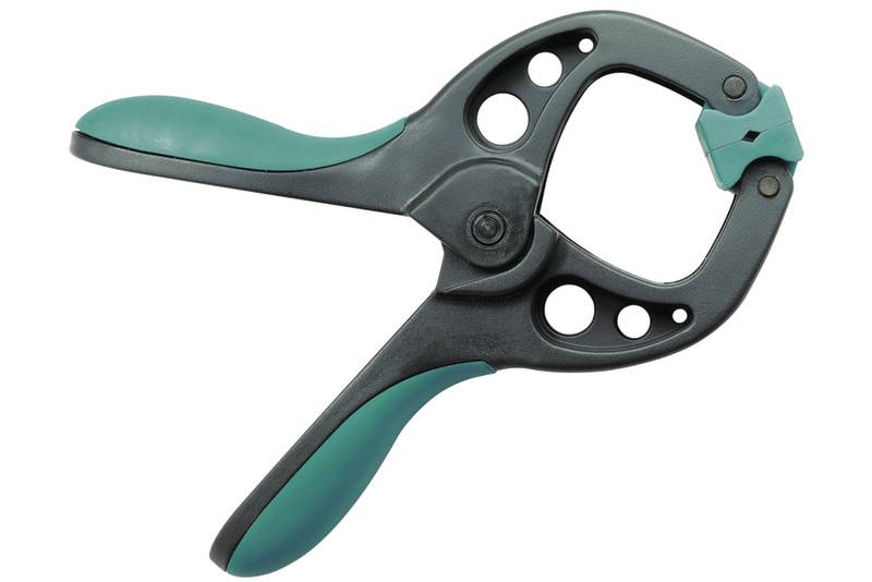 Eine schwarze Spannzange mit grünen Griffen, die zum Halten oder Fixieren von Objekten verwendet wird. Sie hat ein scharnierartiges Design.