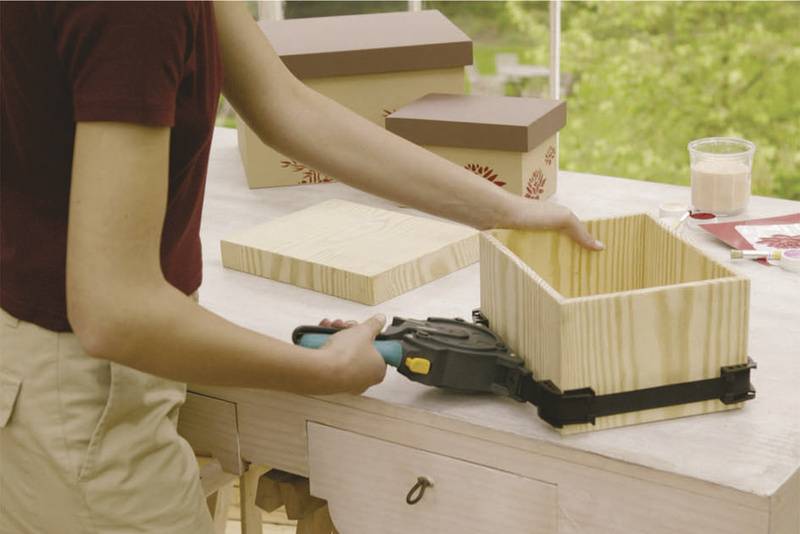 Eine Person benutzt ein Spannwerkzeug, um eine Holzbox zu montieren. Auf dem Tisch befinden sich weitere Holzteile und Boxen.