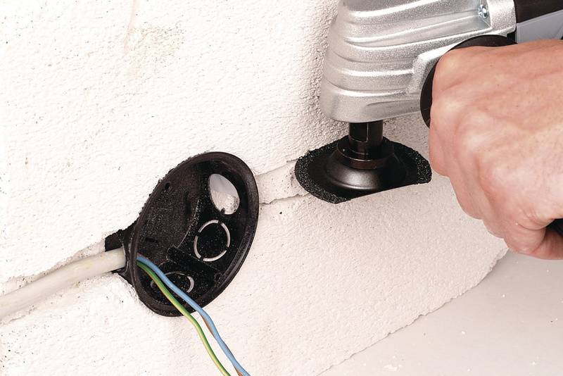 Eine Hand hält ein Werkzeug, das eine runde Öffnung in eine weiße Wand schneidet, neben einer bereits vorhandenen Wandsteckdose.