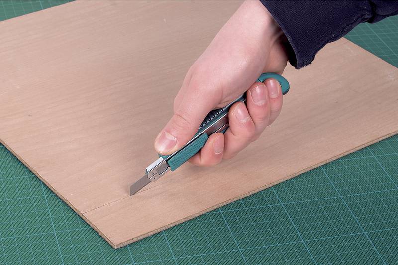 Eine Hand schneidet mit einem Cutter-Messer eine dünne Holzplatte auf einem grünen Schneidematte.