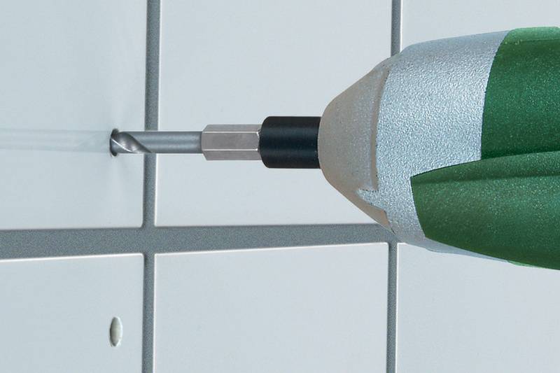 Ein Bohrer durchbohrt eine weiße Fliese in einem Rastermuster. Der Bohrer hat einen grünen Griff.
