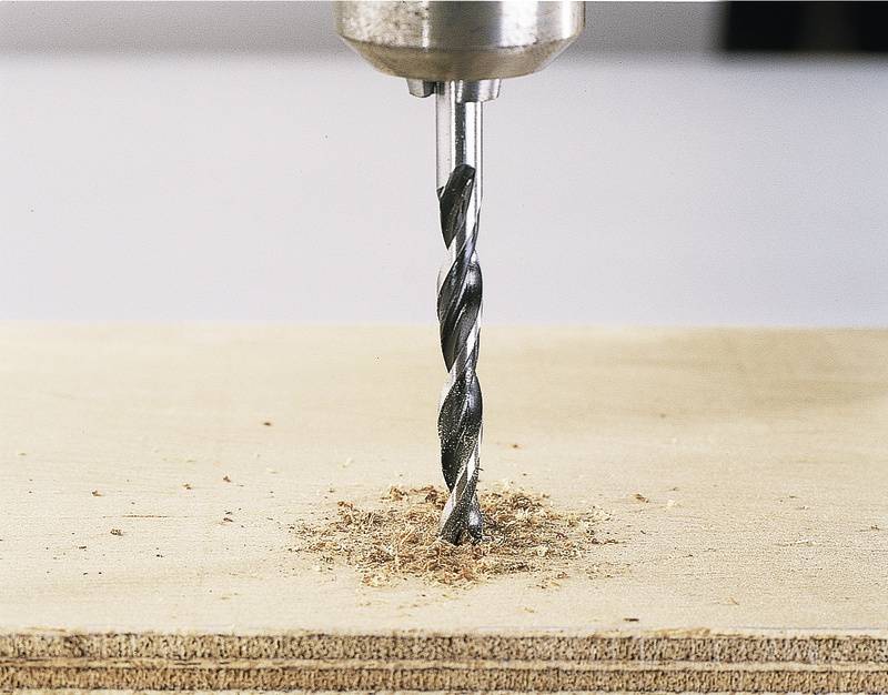 Eine Bohrmaschine bohrt ein Loch in ein Holzbrett. Holzspäne sind um das Loch herum verteilt.