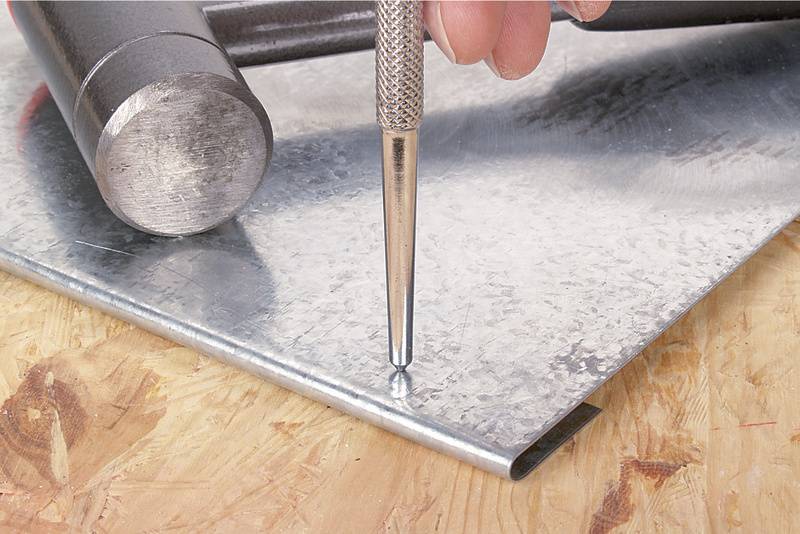 Eine Hand hält einen Körner auf einem Metallblech, bereit zum Ankörnen. Ein Hammer liegt daneben auf der Holzoberfläche.