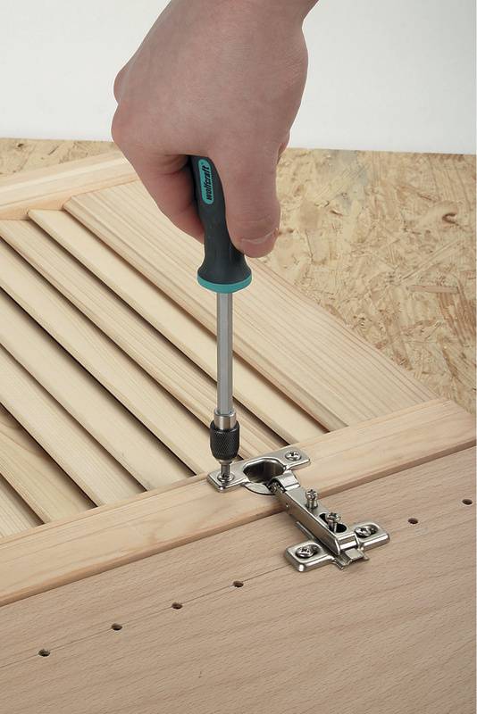 Eine Hand schraubt mit einem Schraubendreher eine Metallscharnier an ein Holzbrett.