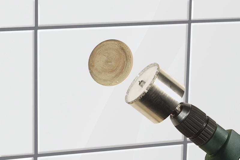 Ein Bohrer mit Lochsägeaufsatz schneidet ein rundes Loch in eine weiße Fliese.