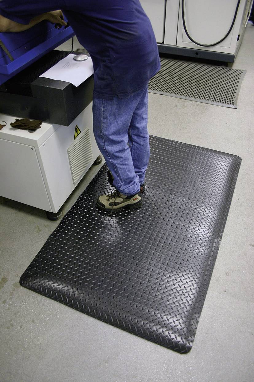 Eine Person steht auf einer schwarzen Anti-Ermüdungsmatte in einer industriellen Umgebung bei einer Maschine.