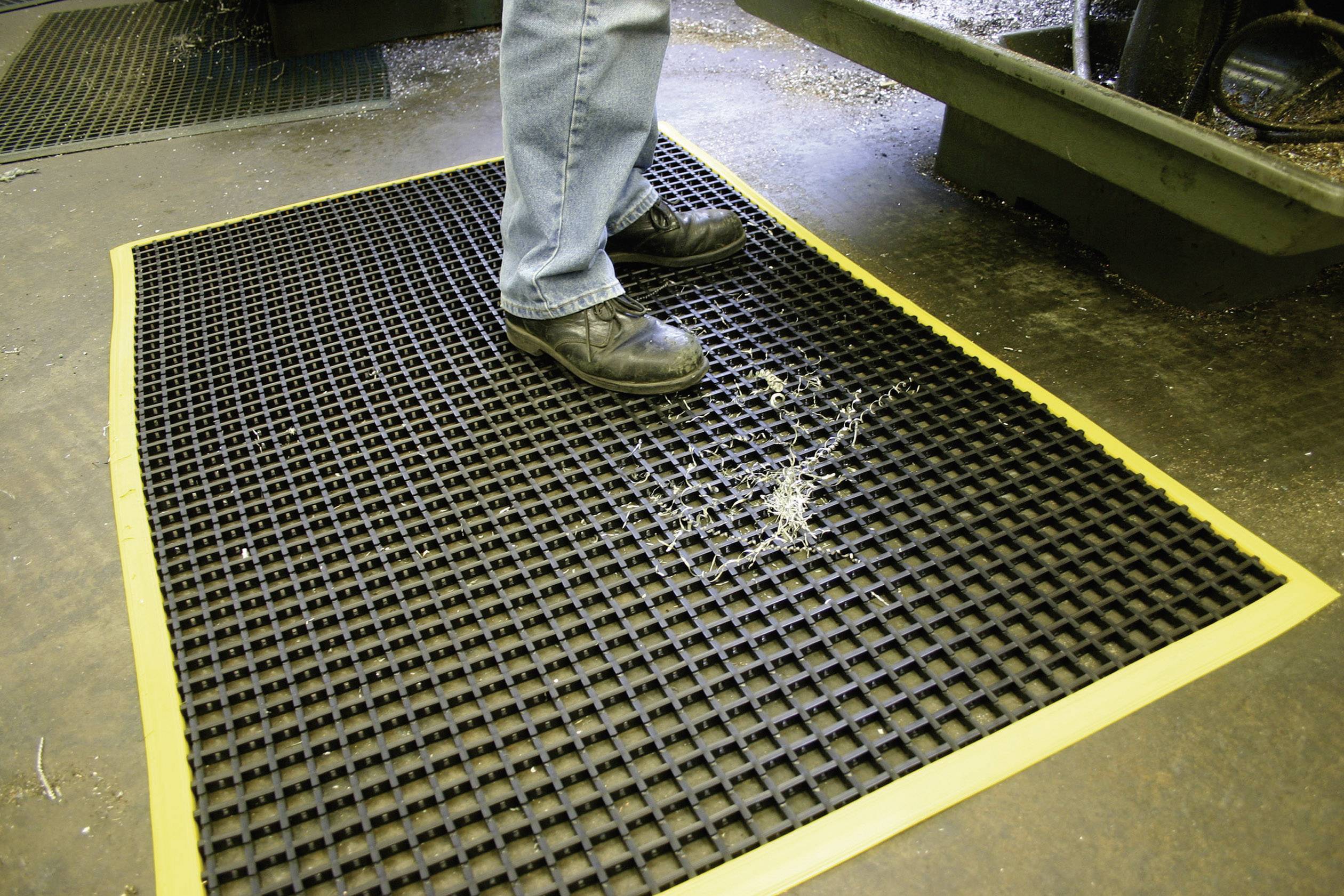 Eine Person steht auf einer rutschfesten Gummimatte mit gelbem Rand, umgeben von Holzspänen. Die Matte verbessert die Arbeitssicherheit.